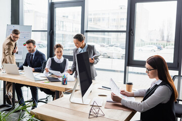 Obraz premium Businesswoman holding papers near coffee, computer and multiethnic colleagues in office