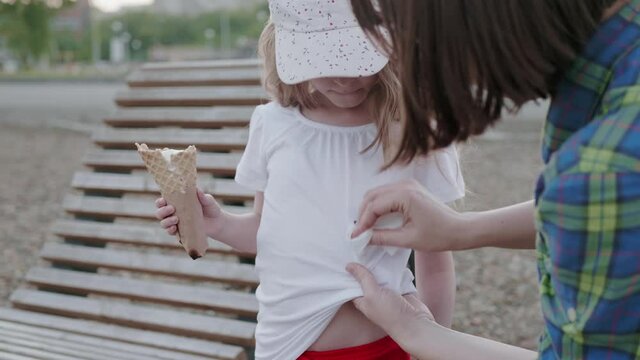 Young unrecognizable mother with napkin wipes chocolate ice cream stains off her little 6 year old daughter's white T-shirt outdoors.