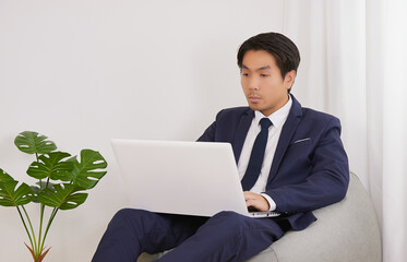 Asian Financial Advisor Using Laptop Computer on Bean Bag in Home Office. Businessman Work at Home for Social Distance or Home Quarantine in Covid 19 or Coronavirus Situation