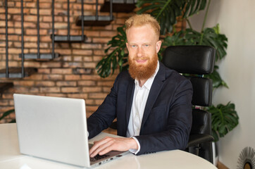 Confident modern red-haired businessman in smart casual suit looks at the camera sitting at the...
