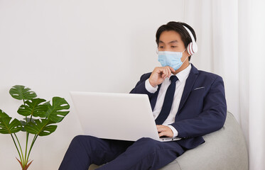 Asian Financial Advisor Wear Mask and Headphone Serious Thinking front Laptop in Home Cafe Office. Businessman Work for Social Distance or Home Quarantine in Covid 19 or Coronavirus Situation