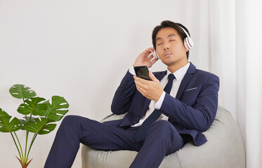 Asian Businessman in Suit Hold Smartphone Wear Headphone for Listen to Music and Sit on Bean Bag in Home Office