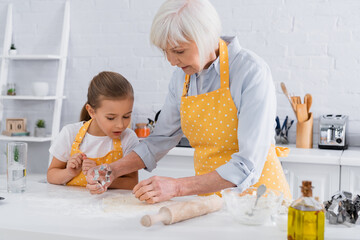 Granny and granddaughter standing near dough and cookie cutter in kitchen