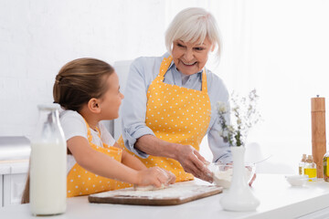 Obraz na płótnie Canvas Cheerful granny holding flour and looking at kid with dough on blurred foreground in kitchen