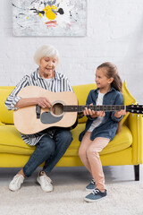 Cheerful grandmother and kid playing acoustic guitar on couch