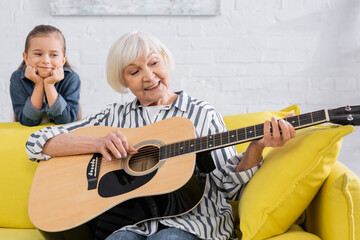 Elderly woman smiling while playing acoustic guitar near blurred kid on couch
