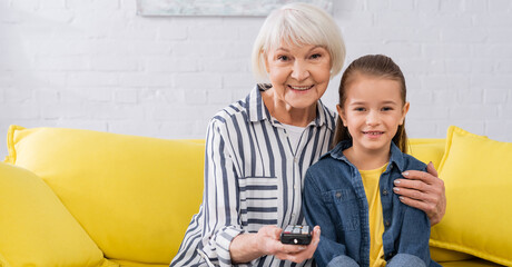 Smiling granny hugging kid while watching tv at home, banner