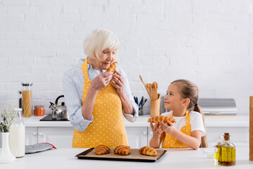 Senior granny smelling croissant near granddaughter in kitchen