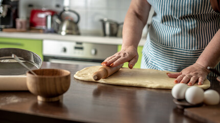 Woman in an apron at home in her kitchen is gently rolling out the dough for making buns. Cooking process of baking