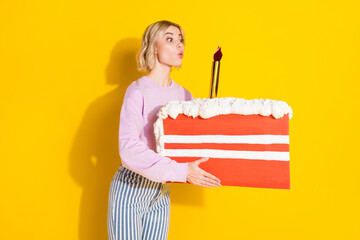 Photo of cheerful nice amazed young lady hold hands big slice cake blow candle isolated on yellow...