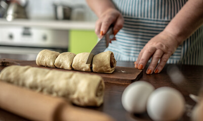 Woman in an apron in the kitchen is gently cutting fresh raw dough with a knife to make delicious homemade cinnamon rolls. Concept of the cooking process of baking