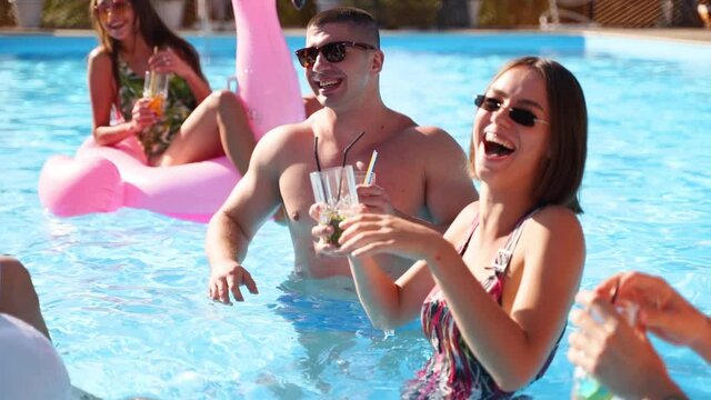 Friends having party with cocktails in holiday villa swimming pool. Happy young people in swimwear dancing, bonding, clubbing with inflatable flamingo, swan, mattress in luxury resort on sunny day.