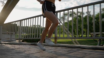 SLOW MOTION CLOSE UP: Female running in green park in small suburban town with tall buildings in the background. Girl jogging across steel bridge leading above heavy traffic highway at golden sunrise - Powered by Adobe