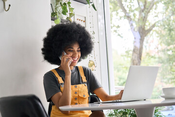 Young happy African American generation z student with curly afro hair sitting in cafe speaking on mobile cell phone, using laptop, working learning remotely indoors in home office. - obrazy, fototapety, plakaty