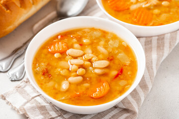 Soup with beans and vegetables in a white bowl on a gray concrete background closeup. Soup made from lentils, chickpeas, white beans, carrots, peppers, and tomatoes. Tasty vegan soup, vegetarian food.