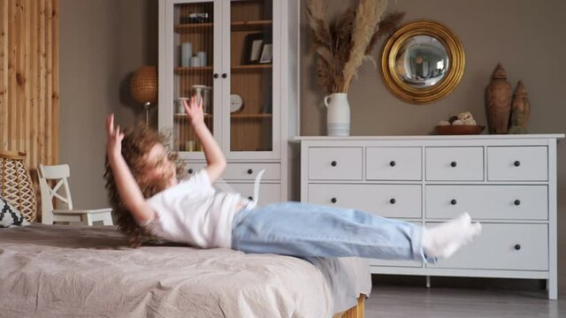 Curly haired preschooler girl in white t-shirt and blue denim jeans jumps and falls back on large grey bed in bedroom at home, slow motion