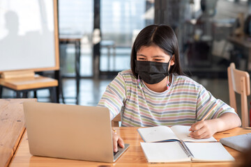Asian woman sitting in room using laptop and taking notes wearing a mask to prevent germs.