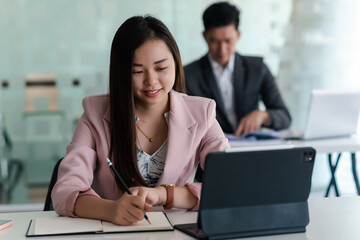 Fototapeta premium Asian businesswoman holding a pencil smiling happy at work using a tablet at the office the back is blurred.