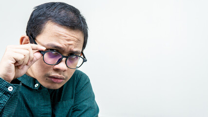 Fototapeta premium Confused face on an Asian Malay man with green shirt concentrate looking at the camera holding pair of spectacles on isolated white background. Checking out the subject in front of him.