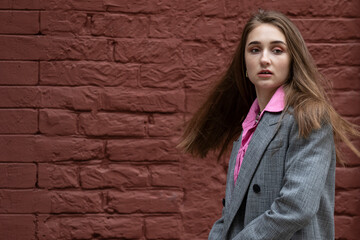 A fashionable girl with hair developing from a sharp turn with her mouth slightly open, dressed in a gray coat and a pink shirt. Teenage girl on a background of a dark brick wall. Copy space.