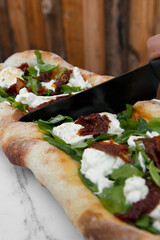 Eating pizza. Closeup view of a hand slicing with a black knife a fresh arugula pizza with Italian burrata cheese and sun dried tomatoes, on the white table. 