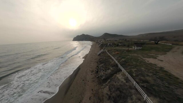 Buggy cars drive along empty sand coastal beach with foaming sea waves past summer constructions and bare trees under cloudy sky aerial fpv view