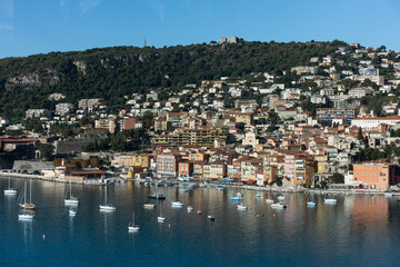 Fototapeta na wymiar Blue sea with small luxury yachts and boats with cute village on a hill at background on sunny summer day. Aerial view of Mediterranean sea in french riviera. High quality photo