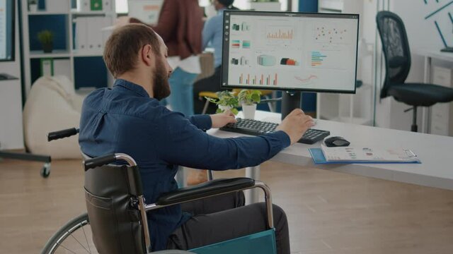 Paralized, immobilized, handicapped marketing manager project sitting in wheelchair in start up business office typing on pc checking reports analysing datas. Diverse team discussing financial project