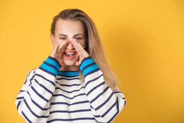 Amused woman yelling, isolated on yellow background. Happy girl holding hands near his mouth
