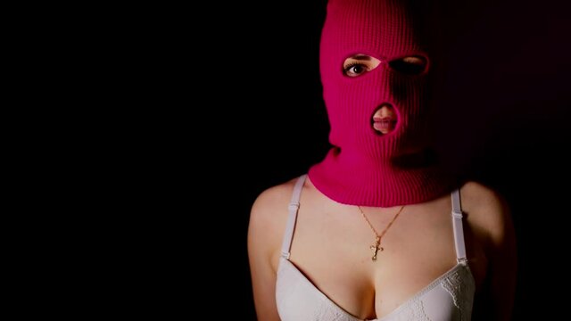 Unrecognizable woman in pink balaclava and white bra stands in dark. Unknown female in mask and lingerie highlighted in dark room.

