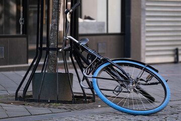 Black bicycle with blue tire is half on the ground and half against a tree in a shopping street, parked carelessly or fallen over. Fallen bicycle