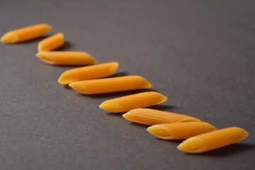Side view of Raw Italian penne pasta pieces scattered on dark background. Some ingredients out of focus. Durum wheat uncooked pasta. Copy space image.
