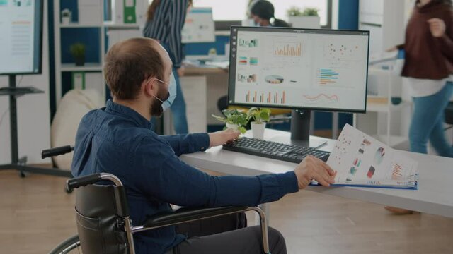Handicapped invalid paralyzed man and teamworkers working in new normal office at financial strategy wearing protective mask. Entrepreneur with disabilities respecting social distance in workspace