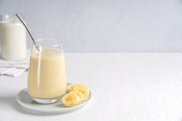 Smoothie or milkshake made of ripe sweet banana fruit blended and mixed with milk served in drinking glass with zero waste metal straw on plate on white wooden table. Image with copy space, horizontal