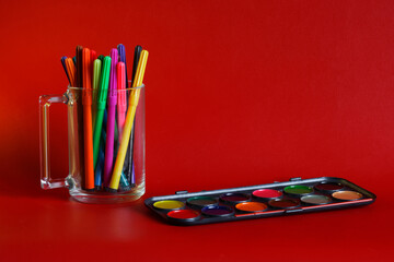 Obraz premium Many multicolored markers in a transparent mug and watercolor paints on a red background. Concept of creativity and learning to draw. Copy space for an inscription