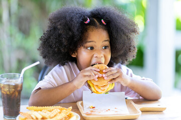 A little curly-haired African-American girl sits on the table with a knife and eats delicious hamburgers and fries. unhealthy food Childhood concepts and eating.