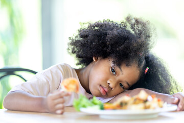 A young, curly haired African American girl sits looking at food. Childhood concepts and healthy...
