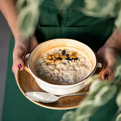 Traditional english breakfast. Woman holds bowl of cereal oatmeal or porridge with milk, raisins and nuts. Close up shot. Soft focus.