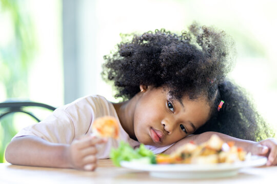 A young, curly haired African American girl sits looking at food. Childhood concepts and healthy eating.