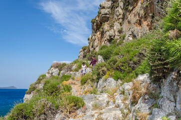People Hike and Climb Across Natural Environment with Bushes and Steep Rock Formations in Monemvasia Island, Peloponnese, Greece. Summer Scenery with Blue Sky and Mediterranean Sea.