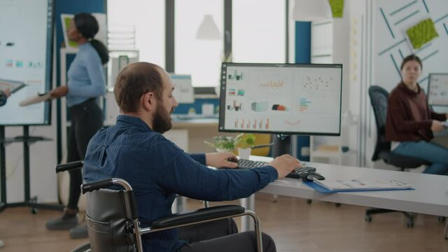Businessman with disabilities asking information to woman worker typing on pc sitting in wheelchair in strat up business office. Colleagues discussing about financial project checking statistics datas
