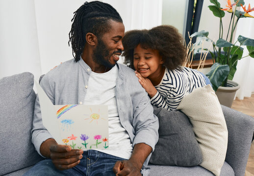 Father-daughter relationship. Dad is happy to receive a greeting card with a child's drawing from his daughter to father's day, sitting on a sofa at home. Happy family