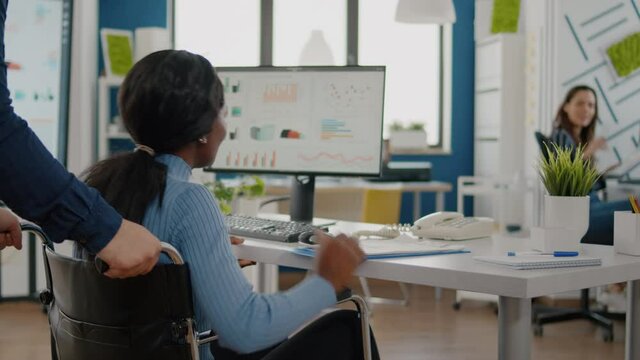 Businessman helping his disabled black colleague entering in company office pushing wheelchair near desk. African handicapped paralysed woman greetting colleagues starting working at financial project