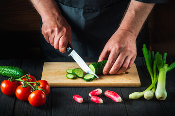Chef or cook cuts a green cucumber on a restaurant kitchen cutting board for salad. Vegetable diet or snack idea