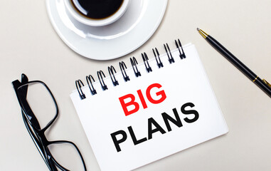 Glasses, a white cup of coffee, a white notebook with the words BIG PLANS and a ballpoint pen lie on a light background. Flat lay. View from above.