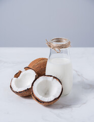 Glass vegan milk bottle on white marble table with half coconuts. Front view and copy space