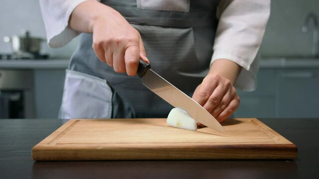 Female hands slice white onion by knife on wooden board on kitchen background