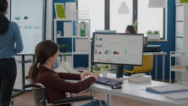 Paralysed financial expert wearing face mask sitting in wheelchair analysing economy statistics of company in new normal business office typing on computer. Handicapped woman using modern technology