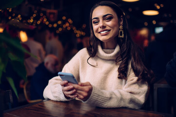 Cheerful woman with evening makeup and hairstyle using phone at cafe