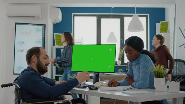 Diverse team working together with financial graphs looking at green screen, mock up, chroma key isolated desktop, black woman and paralysed coworker in wheelchair discussing solution for project